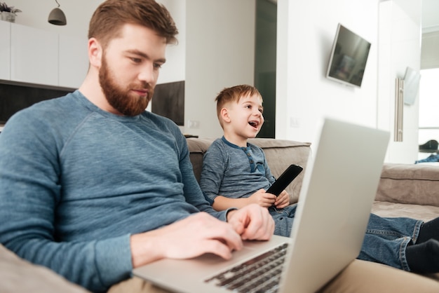 Foto bild des jungen vaters mit laptop-computer, während sein kleiner sohn fernsieht.