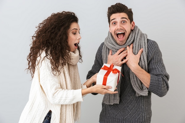 Bild des kaukasischen Mannes und der Frau, die sich freuen, während sie mit Geschenkbox stehen, lokalisiert über graue Wand