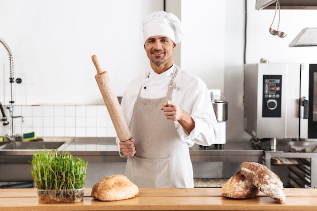 Bild des professionellen Mannbäckers in der weißen Uniform lächelnd, während an der Bäckerei mit Brot auf Tisch stehend