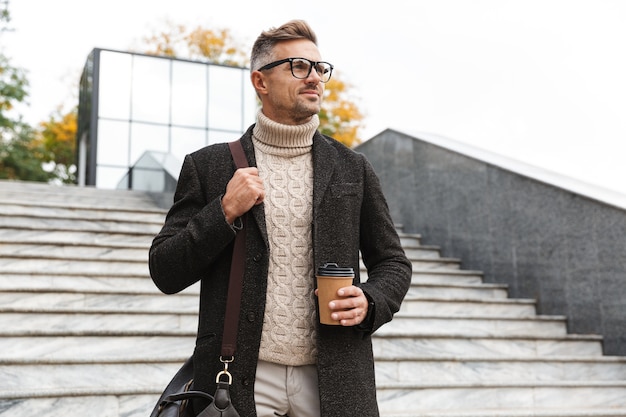 Bild des reifen Mannes 30s, der Brillen trägt, durch Stadtstraße geht und Kaffee zum Mitnehmen hält