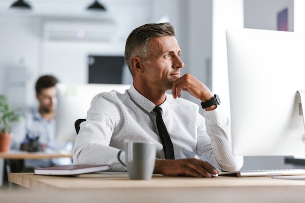 Bild des schönen Geschäftsmannes 30s, der weißes Hemd und Krawatte trägt, die am Schreibtisch im Büro durch Computer sitzen und beiseite schauen