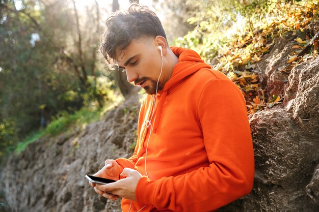 Bild des schönen jungen sportfitnessmannläufers draußen im park, der musik mit kopfhörern unter verwendung des mobiltelefons hört.