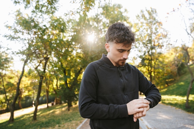 Bild des schönen jungen Sportfitnessmannläufers draußen im Park, der Uhr schaut.