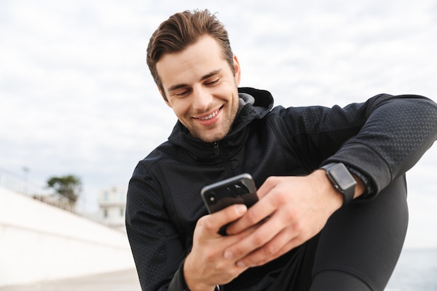 Bild des sportlichen Mannes 30s in der schwarzen Sportbekleidung, unter Verwendung des Smartphones beim Sitzen auf der Promenade am Meer