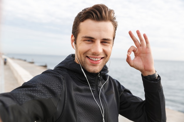 Bild des zufriedenen Sportlers 30s in der schwarzen Sportbekleidung und in den Kopfhörern, die Selfie-Foto auf Handy während des Gehens am Meer machen