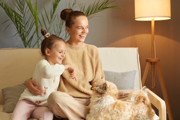 Bild einer äußerst glücklichen Frau mit Brötchenfrisur und kleiner Tochter, die mit Pekingese-Hund spielt, während sie auf Husten sitzt und glücklich lacht und sehr gute Laune hat