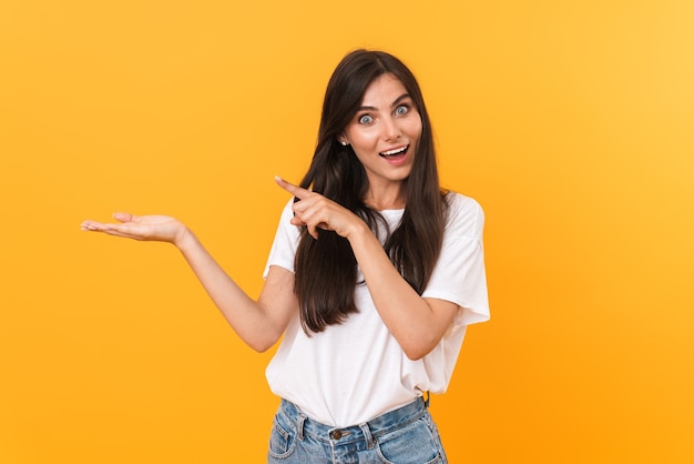 Bild einer aufgeregten brünetten Frau mit langen Haaren, die lächelt und mit den Fingern auf das Exemplar auf ihrer Handfläche zeigt, isoliert über gelber Wand