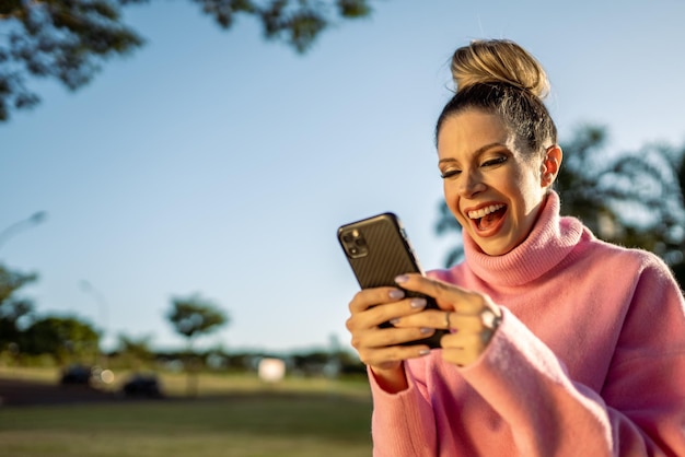 Bild einer aufgeregten jungen blonden lateinamerikanischen Frau, die glücklich ist, ein Mobiltelefon auf Parkhintergrund zu benutzen