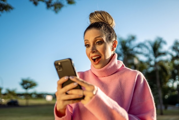 Bild einer aufgeregten jungen blonden lateinamerikanischen Frau, die mit einem Mobiltelefon auf Parkhintergrund überrascht wurde