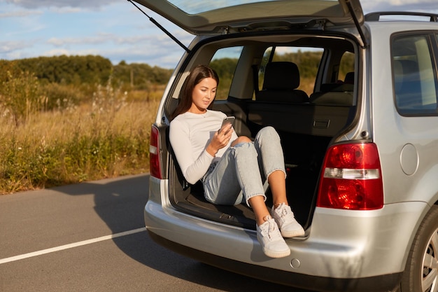 Bild einer dunkelhaarigen jungen erwachsenen Frau, die im Kofferraum eines Autos sitzt und ein Handy benutzt, das legere Kleidung trägt und im Internet surft, während sie soziale Netzwerke überprüft