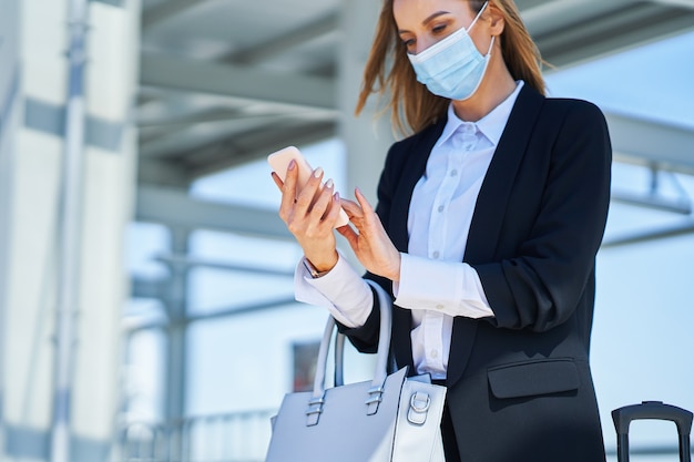 Bild einer eleganten Frau mit Schutzmaske, die mit Tasche und Koffer im Bahnhof spaziert