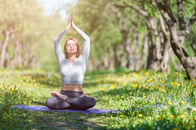 Bild einer Frau, die Yoga mit erhobenen Armen macht, die an einem Sommertag im Lotussitz auf einem blauen Teppich im Wald sitzt?