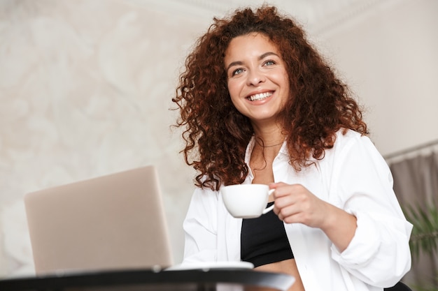 Bild einer fröhlichen, glücklichen lächelnden jungen Frau, die im weißen Hemd zuhause im Haupthotel unter Verwendung des trinkenden Kaffees der Laptop-Computer gekleidet wird.