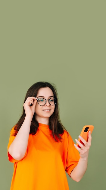 Bild einer fröhlichen jungen Frau in einem orangefarbenen T-Shirt mit einem modernen Smartphone in einem leeren grünen Raum