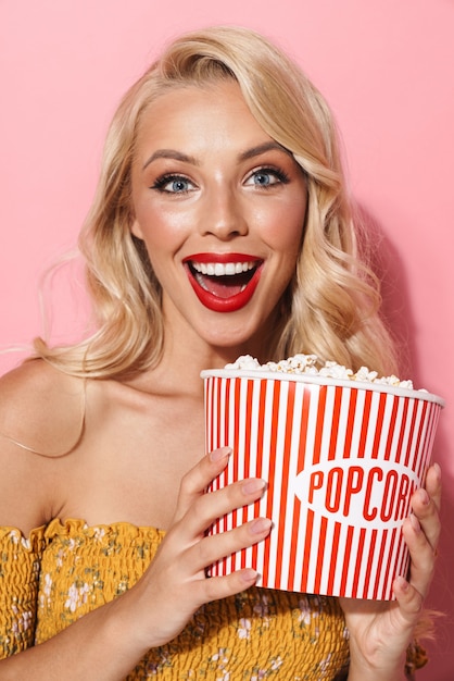 Bild einer glamourösen jungen Frau mit rotem Lippenstift, die lächelt und Popcorn-Eimer hält, isoliert über rosa Wand