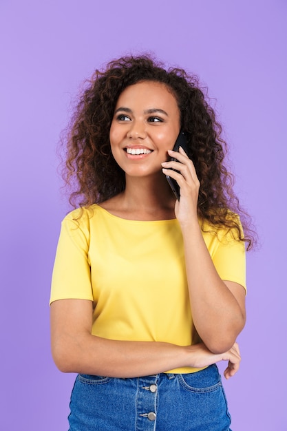 Bild einer glücklichen Afroamerikanerin mit lockigem Haar, die auf dem Smartphone spricht, isoliert über der violetten Wand?