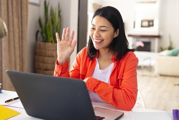 Bild einer glücklichen biracial Frau, die am Schreibtisch sitzt und während eines Videoanrufs auf dem Laptop zu Hause winkt, Platz zum Kopieren. Arbeiten von zu Hause aus, Technologie, Kommunikation und Lifestyle-Konzept.