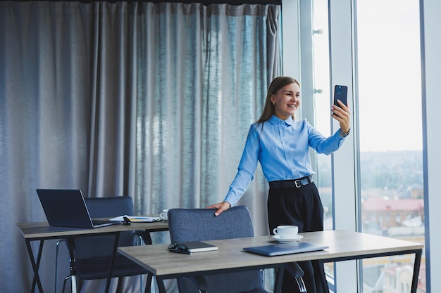 Bild einer glücklichen jungen Frau in einer Jacke, die lächelt und an einem Laptop arbeitet und in einem modernen Büro mit großen Fenstern telefoniert Fernarbeit