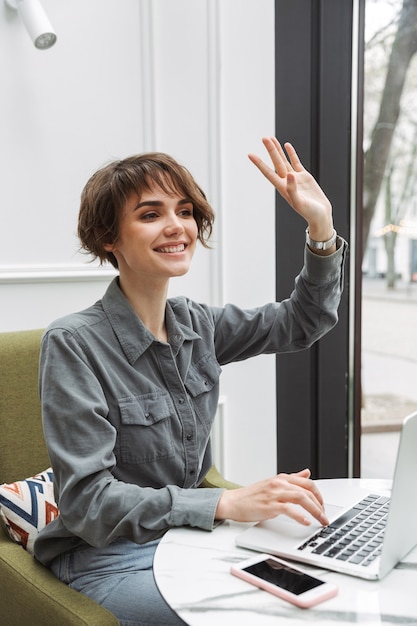 Bild einer glücklichen jungen hübschen Geschäftsstudentenfrau, die im Café zuhause sitzt und Laptop-Computer coworking verwendet, die zu Freunden wellenartig bewegt.