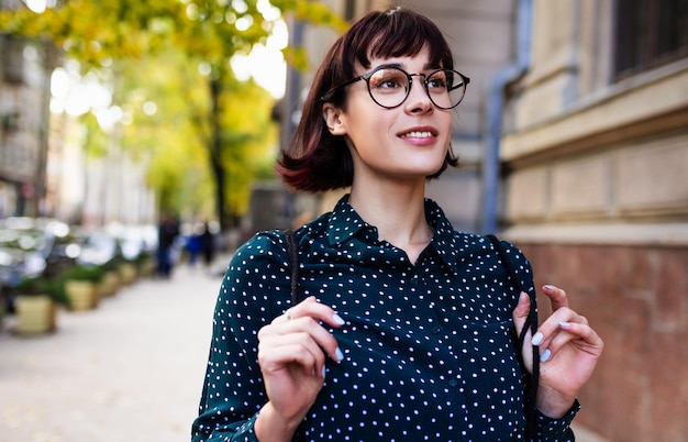 Bild einer intelligenten, schönen jungen Frau, die eine transparente Brille mit Rucksack trägt, die aufs College geht Junge Studentin, die neben dem Gebäude auf der Straße zu Fuß ist