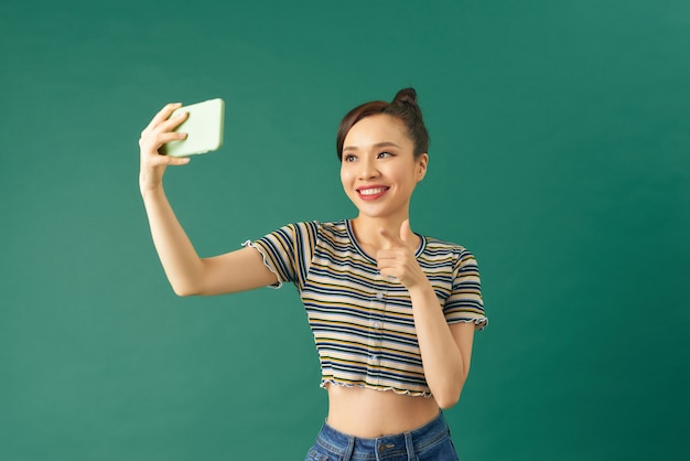 Bild einer jungen asiatischen Frau, die lächelt und ein Selfie-Foto auf dem Handy macht, isoliert auf grünem Hintergrund