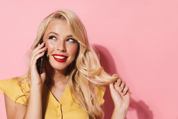 Bild einer jungen Frau mit langen blonden Haaren, die lächelt und auf dem Handy spricht, isoliert über rosa Wand