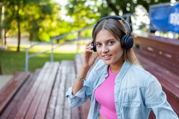 Bild einer jungen stilvollen Frau in modischer Jugendkleidung, die Musik mit Kopfhörern hört, während sie auf den Ständen im Freien sitzt