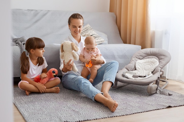 Bild einer kaukasischen dunkelhaarigen Frau in weißem Hemd und Jeans, die mit ihren Kindern spielt und ihrem kleinen Mädchen, das auf dem Boden neben dem Sofa sitzt, ein weiches Hundespielzeug zeigt