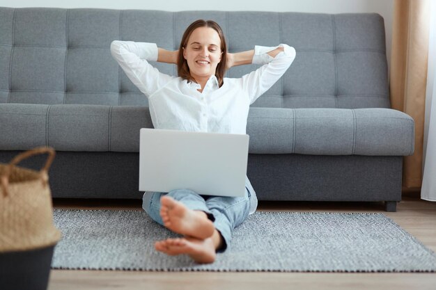 Bild einer kaukasischen dunkelhaarigen Frau mit weißem Hemd und Jeans, die auf dem Boden sitzt und am Laptop arbeitet, das Display mit einem zahnigen Lächeln betrachtet, die Hände hebt und mit ihrer Arbeit zufrieden ist.