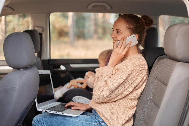 Bild einer lächelnden, entzückten kaukasischen Frau, die einen beigen Pullover trägt und mit ihrem Baby auf dem Rücksitz sitzt und ein Smartphone spricht, das einen PC-Computer auf den Knien hält