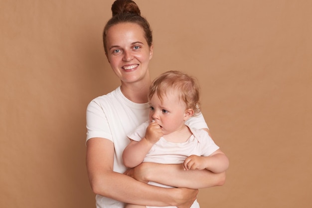Bild einer lächelnden, fröhlichen Frau mit Brötchenfrisur, die mit ihrem Kleinkind posiert, das in die Kamera blickt, während sie ihre kleine Tochter isoliert vor braunem Hintergrund hält