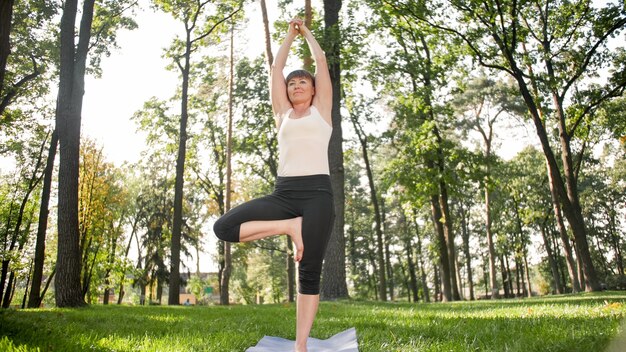 Bild einer lächelnden glücklichen Frau mittleren Alters, die meditiert und Yoga-Übungen auf Gras im Wald macht. Frau, die sich um ihre körperliche und geistige Gesundheit kümmert, während sie Fitness und Stretching im Park übt