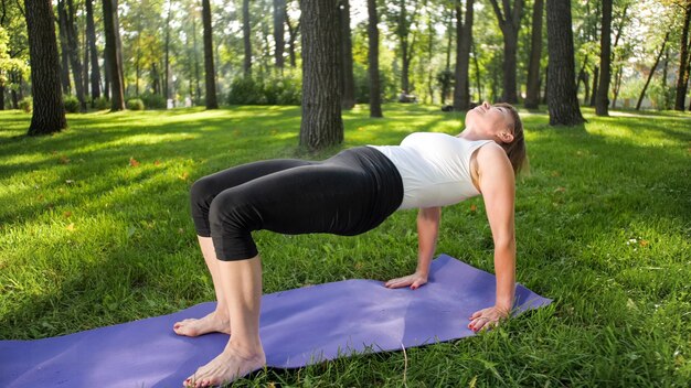 Bild einer lächelnden glücklichen Frau mittleren Alters, die meditiert und Yoga-Übungen auf Gras im Wald macht. Frau, die sich um ihre körperliche und geistige Gesundheit kümmert, während sie Fitness und Stretching im Park übt