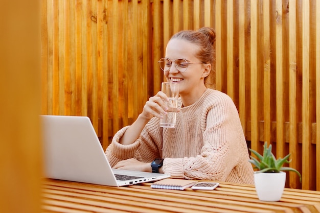 Bild einer lächelnden hübschen kaukasischen Frau mit Brötchenfrisur, die einen beigen Pullover trägt, der an einem tragbaren Computer vor einer Holzwand arbeitet und auf das Display Trinkwasser aus Glas blickt