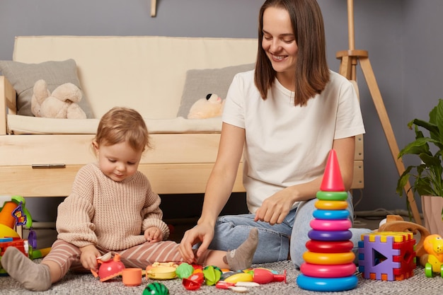 Bild einer lächelnden kaukasischen lächelnden Frau und Tochter, die zusammen spielen, während sie zu Hause auf dem Boden sitzen Aktivitäten mit Kindern in der frühkindlichen Entwicklung