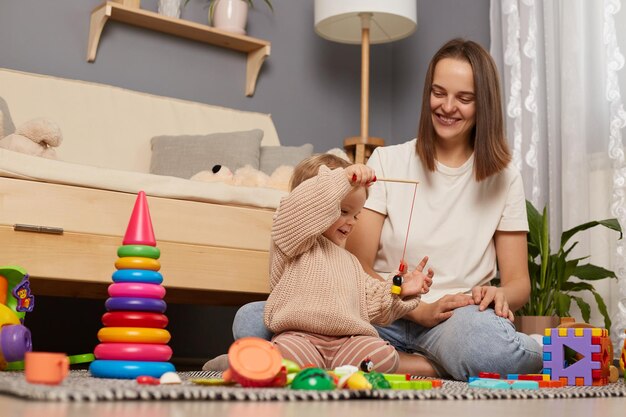 Bild einer lächelnden Mutter, die mit einem Kleinkindmädchen spielt, während sie auf einem Teppich im Kinderzimmer neben dem Sofa sitzt, ein weibliches Kind, das eine Angelrute mit einem Magneten hält und frühe Entwicklungsspiele spielt