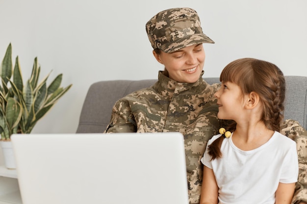 Bild einer lächelnden, positiven Frau in Tarnuniform und Mütze Militärfrau, die nach der Armee nach Hause zurückkehrt und Zeit mit ihrer Tochter verbringt, die sitzt und etwas auf dem Laptop anschaut