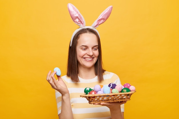 Bild einer lächelnden, schönen, fröhlichen Europäerin mit Hasenohren, die Ostereier hält und wegschaut und die Osterfeier genießt, die isoliert auf gelbem Hintergrund posiert