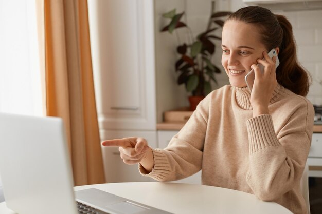 Bild einer optimistisch lächelnden Frau mit beigem Pullover, die in der Küche sitzt und online am Laptop arbeitet und per Handy mit dem Finger auf das Notebook-Display zeigt