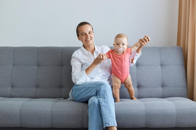 Bild einer positiv optimistischen Frau mit weißem Hemd und Jeans, die mit ihrer kleinen Tochter auf dem Sofa sitzt, Baby in rosa Bodysuit, das mit der Hilfe der Mutter steht.