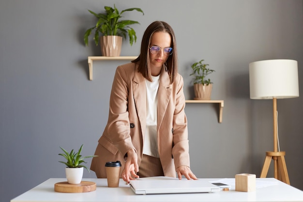 Bild einer ruhigen, ernsthaften, konzentrierten, attraktiven jungen erwachsenen kaukasischen Frau, die eine beige Jacke trägt, die im Büro posiert und bereit ist, mit der Arbeit zu beginnen, öffnet ihren Laptop