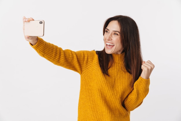 Bild einer schönen brünetten erwachsenen Frau mit Pullover, die lächelt und ein Selfie-Foto auf dem Handy macht, isoliert auf weiß