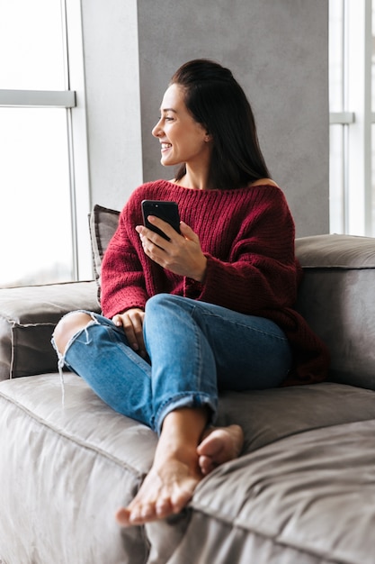 Bild einer schönen Frau drinnen im Haus auf Sofa unter Verwendung des Mobiltelefons.