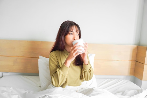 Bild einer schönen jungen asiatischen Frau, die ein Sweatshirt trägt, lächelt und Tee auf dem Bett in einer weißen Wohnung trinkt