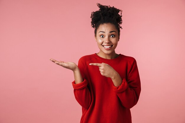 Bild einer schönen jungen aufgeregten glücklichen emotionalen afrikanischen Frau, die über rosa Wand zeigt aufzeigt.