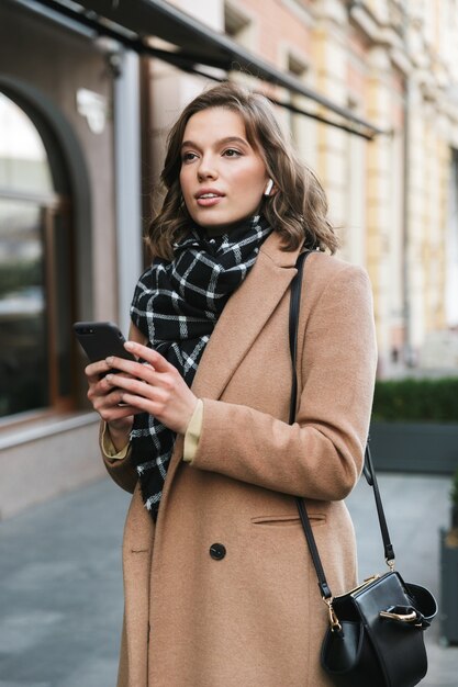 Bild einer schönen jungen Frau im Freien, die durch Straße geht, die Handy hörend Musik mit Kopfhörern hört.