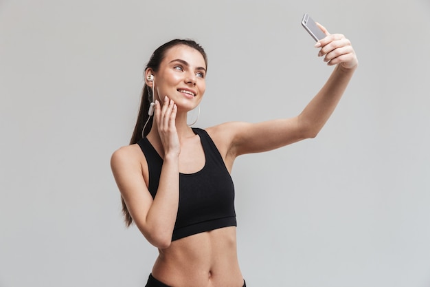 Bild einer schönen jungen Sport-Fitness-Frau, die über graue Wand isolierte Musik des Handys hört, macht ein Selfie.