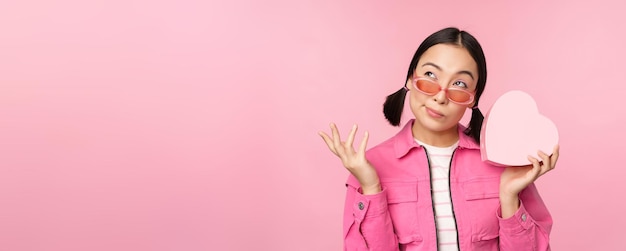 Bild einer stilvollen asiatischen Freundin mit Sonnenbrille, die errät, was sich in der herzförmigen Geschenkbox befindet, die über rosa Hintergrund steht