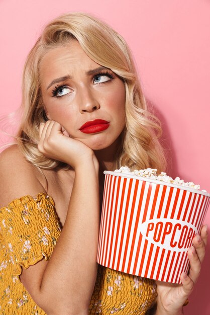 Bild einer traurigen jungen Frau mit rotem Lippenstift, die Popcorn-Eimer isoliert über rosa Wand hält