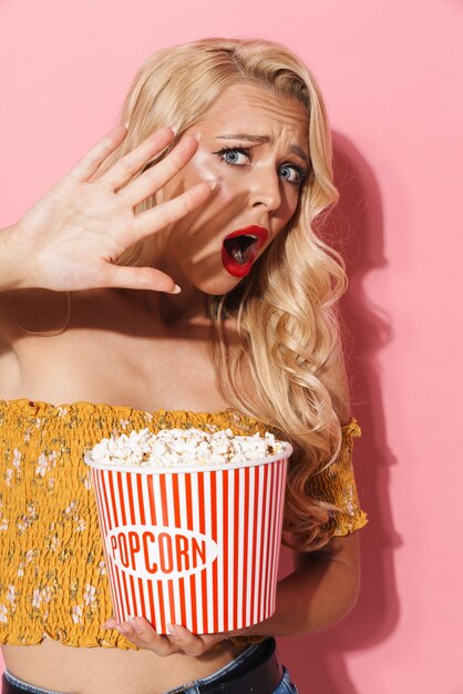 Bild einer verängstigten jungen Frau mit rotem Lippenstift, die schreit und Popcorn-Eimer hält, isoliert über rosa Wand